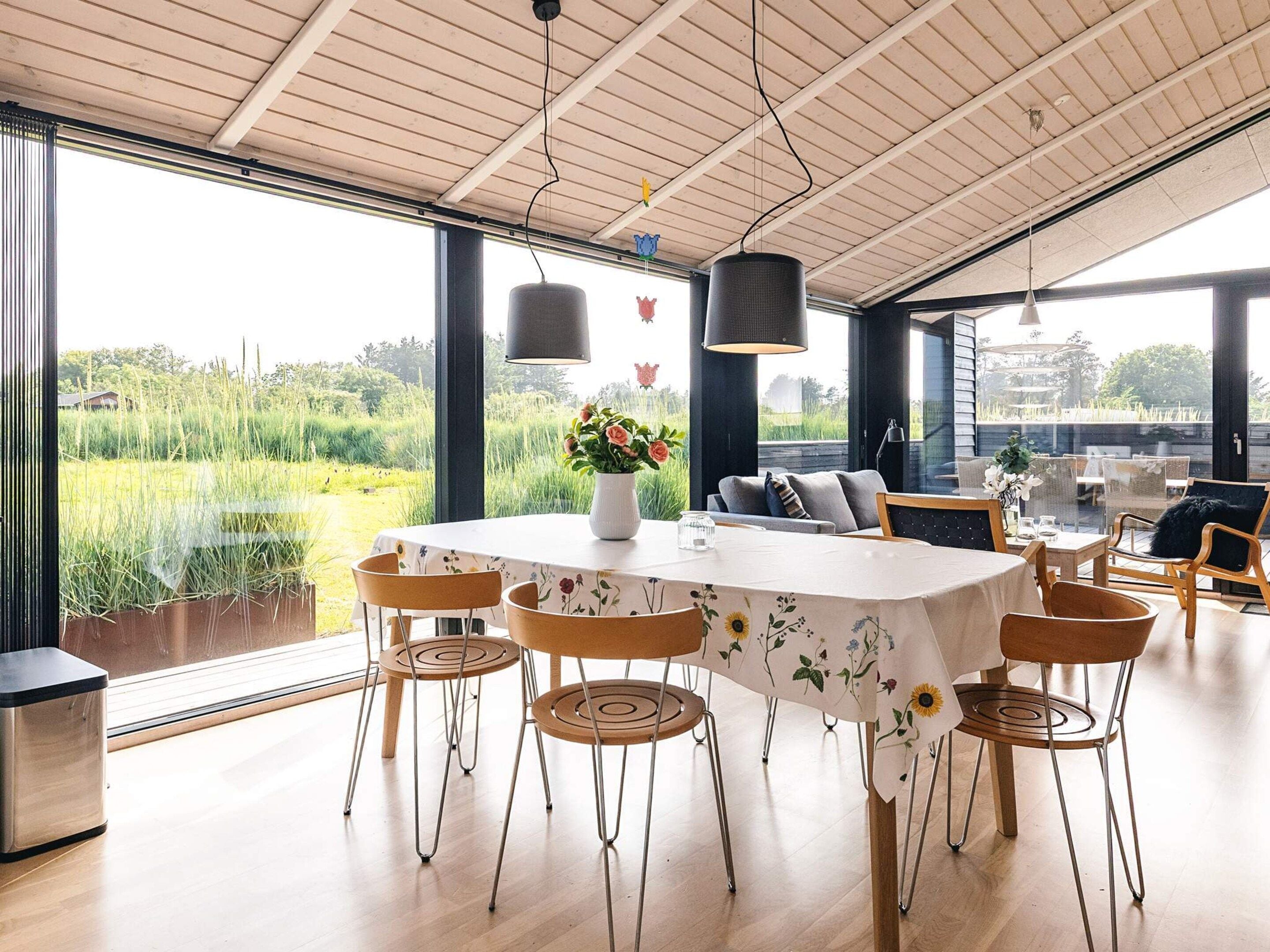 A dining area with a round table, six chairs, a vase with flowers, and a view of a field through large windows.
