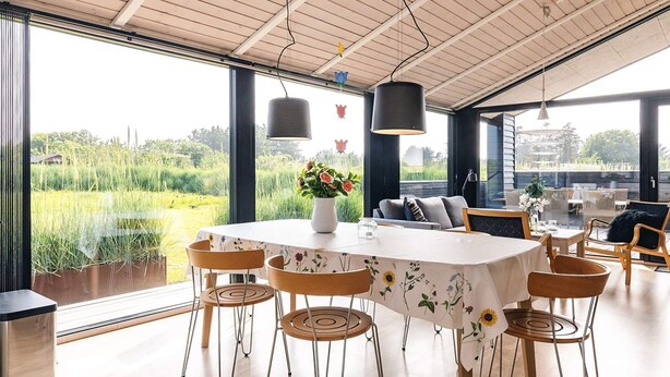 A dining area with a round table, six chairs, a vase with flowers, and a view of a field through large windows. - 5 Star Holiday Home in Hirtshals (Hirtshals)