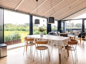 A dining area with a round table, six chairs, a vase with flowers, and a view of a field through large windows. - 5 Star Holiday Home in Hirtshals (Hirtshals)