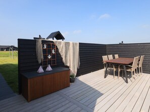 A wooden deck with a table and chairs, a small bench with cushions, and a decorative wall with a window and hanging ornaments.