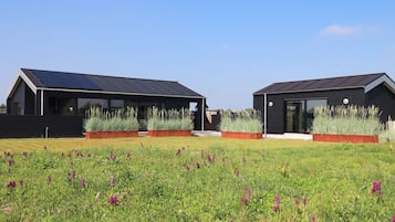 Two modern, dark-colored buildings with solar panels, surrounded by a grassy field with purple flowers.