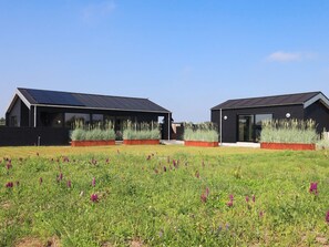 Two modern, dark-colored buildings with solar panels, surrounded by a grassy field with purple flowers.