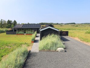 A modern house with a gravel driveway, a wooden structure adjacent to it, and a grassy field in the background. - 5 Star Holiday Home in Hirtshals (Hirtshals)