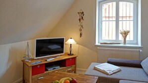 A cozy room with a television on a wooden stand, a small table with fruits, a window with a red valance, and a radiator. - Apartment in Lancken-granitz (Lancken-Granitz)