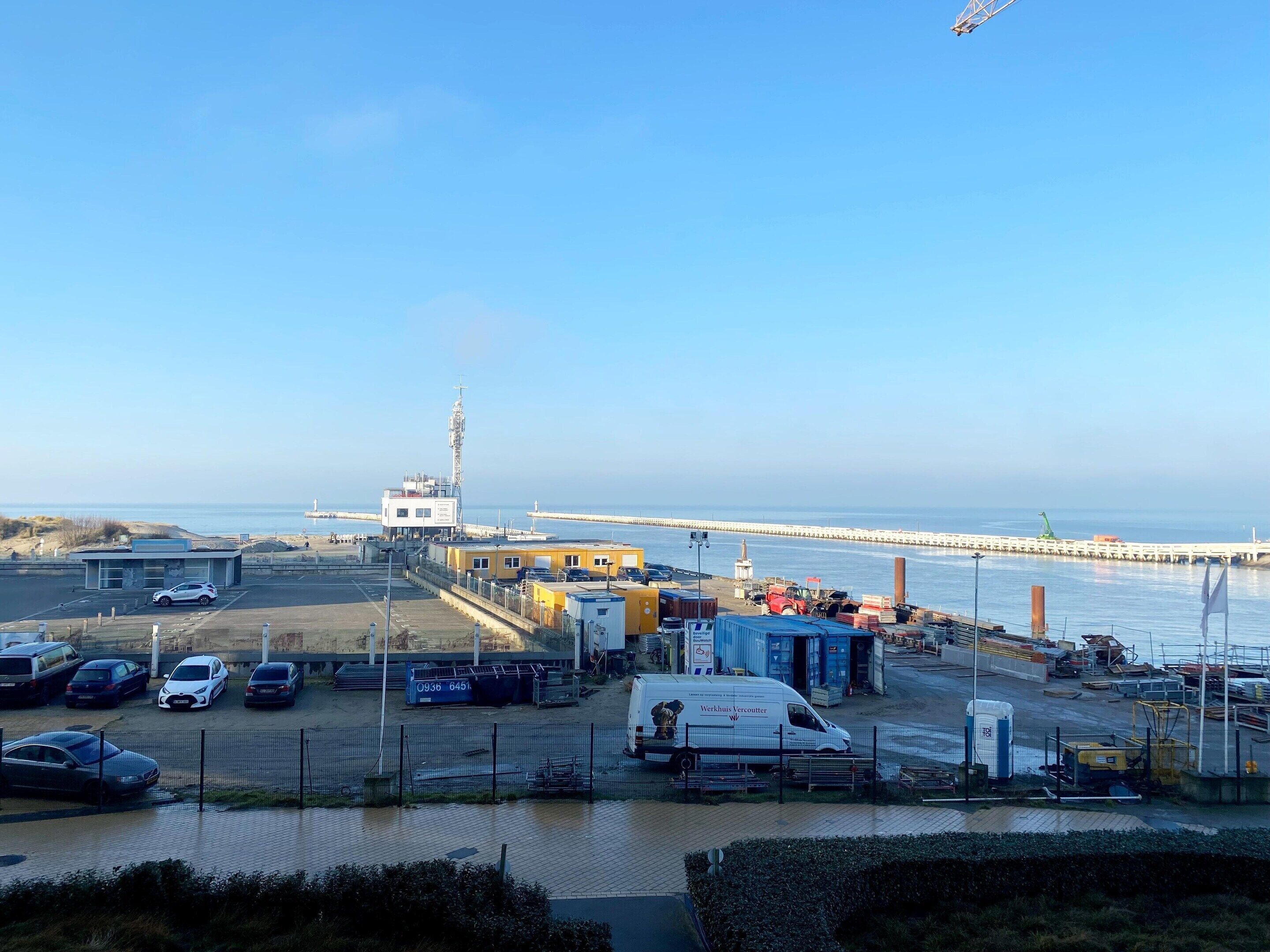 A coastal industrial area with a pier extending into the water, a white van, and several parked cars.