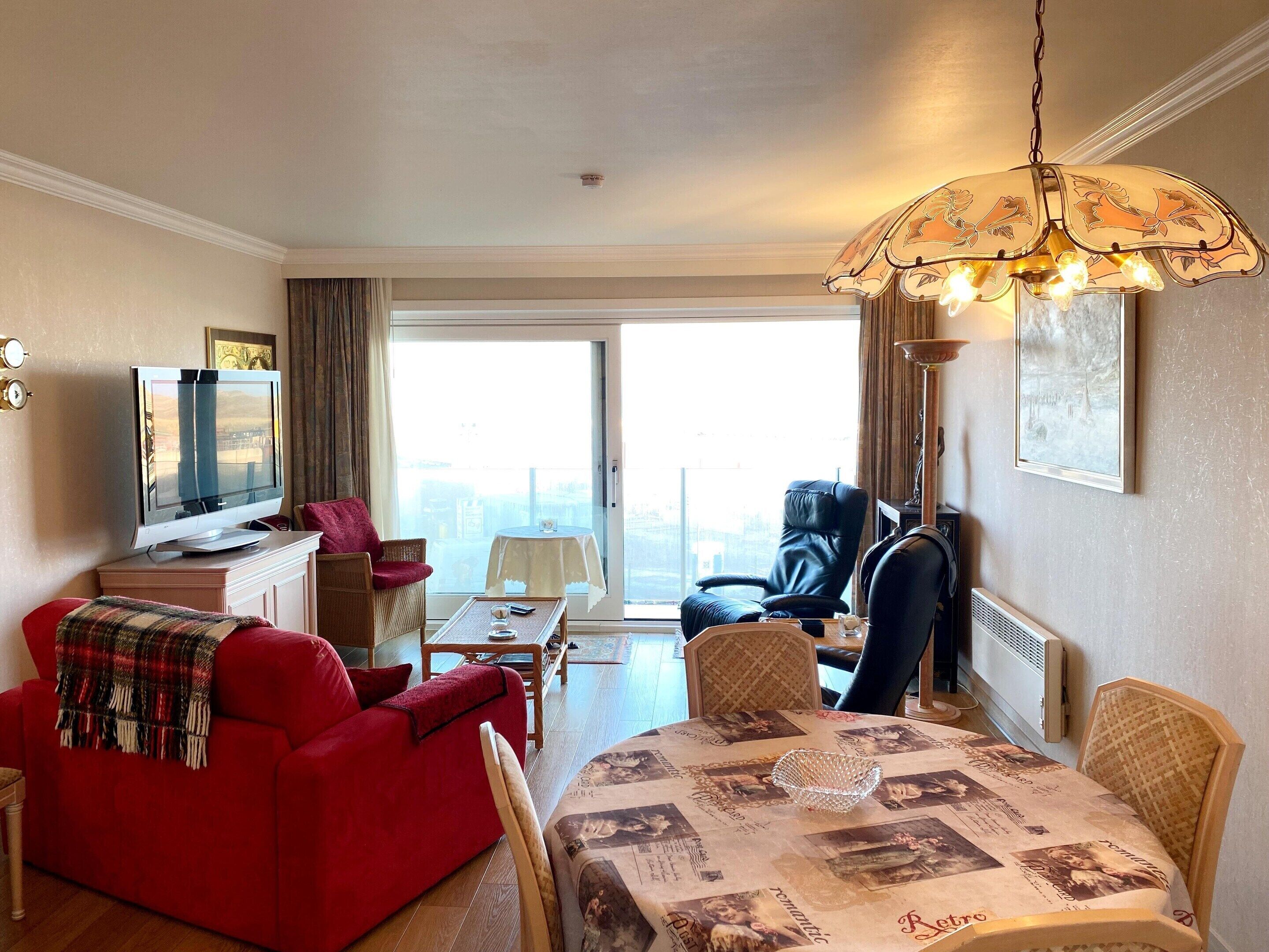 Een hotelkamer met een rode bank, een eettafel met een vintage tafelkleed, een zwarte fauteuil, een televisie en een groot raam met uitzicht op de stad.