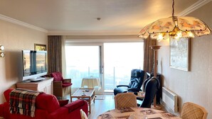 A hotel room with a red sofa, a dining table with vintage tablecloth, a black armchair, a television, and a large window with a city view. - Apartment in Nieuwpoort Near Marina (Nieuwpoort)