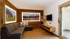 A modern living room with a sectional sofa, wooden paneling, a large window with a mountain view, a mounted TV, and a sliding glass door. - Holiday Home Hutterwirtshof (Moso in Passiria)