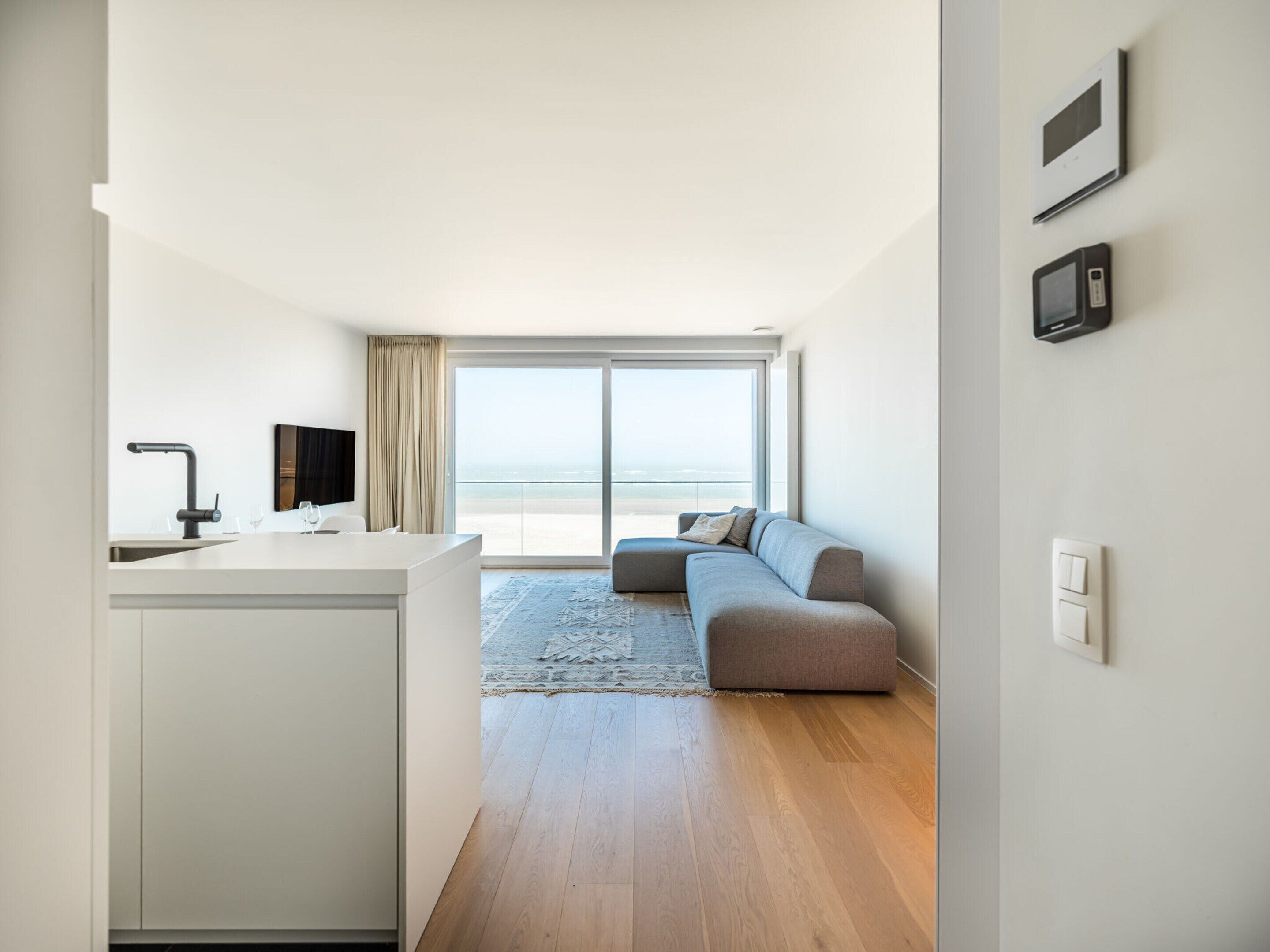 Ein moderner Wohnraum mit Küche, einem großen Fenster mit Aussicht, einem grauen Sofa und einem Flachbildfernseher.