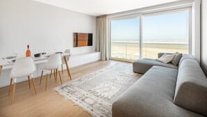 A modern living room with a large window overlooking a beach, a dining area with white chairs, and a grey sofa.