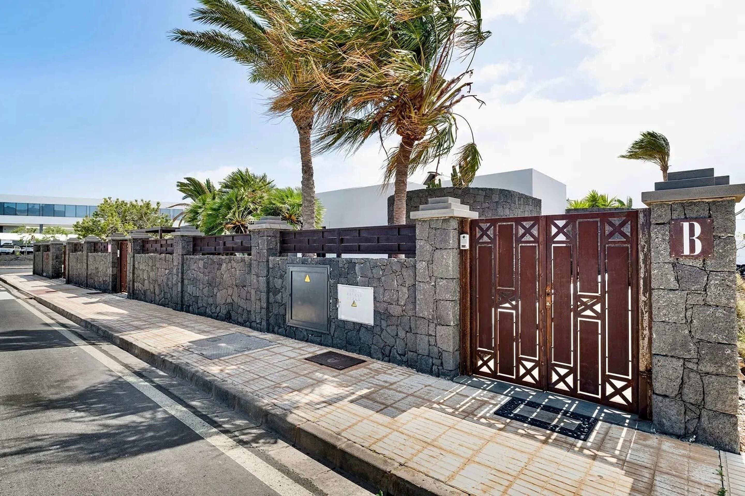 A gated entrance with a stone wall, palm trees, and a modern building in the background.