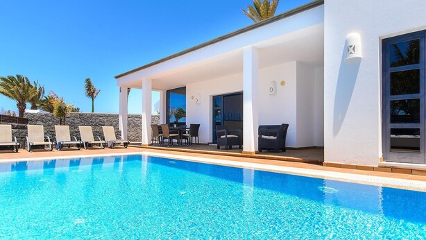 Un área de piscina moderna con una piscina azul cristalina, sillas de descanso y un porche cubierto con mobiliario de exterior.