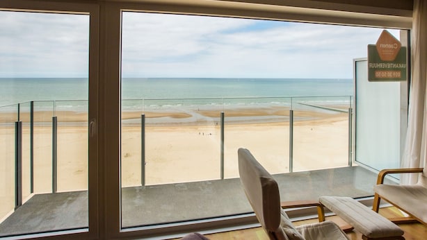 A room with a large window overlooking a beach, a chair, and a bench. - Lovely Apartment for a Family (Middelkerke)