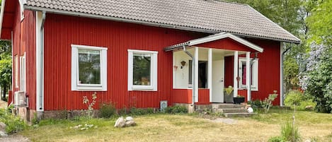 A red wooden house with a tiled roof, a porch, and a small garden.