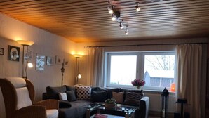 A cozy living room with a wooden ceiling, a large window with curtains, a sofa, a chair, and a coffee table. - Kassler Comfortable Holiday Residence (Feldberg)