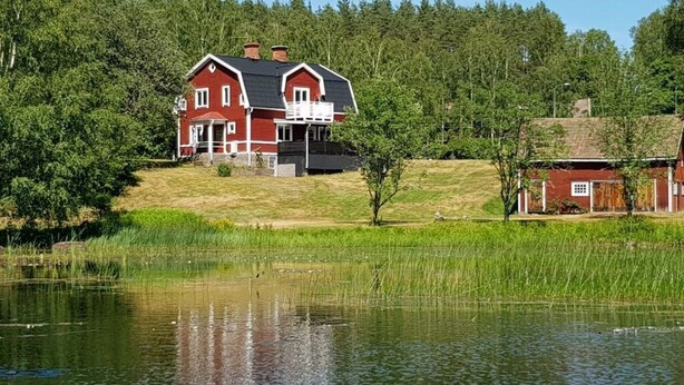 A red house with a balcony, surrounded by a pond and trees. - Country House Dalavagen 8 (Lönneberga)
