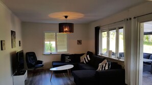 A modern living room with a black sofa, a white coffee table, and a TV. There are windows with blinds, a ceiling light, and a view of the outdoors. - Country House Dalavagen 8 (Lönneberga)