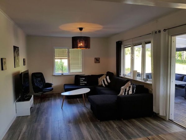 Un salon moderne avec un canapé noir, une table basse blanche et un téléviseur. Il y a des fenêtres avec des stores, un luminaire au plafond et une vue sur l’extérieur.
