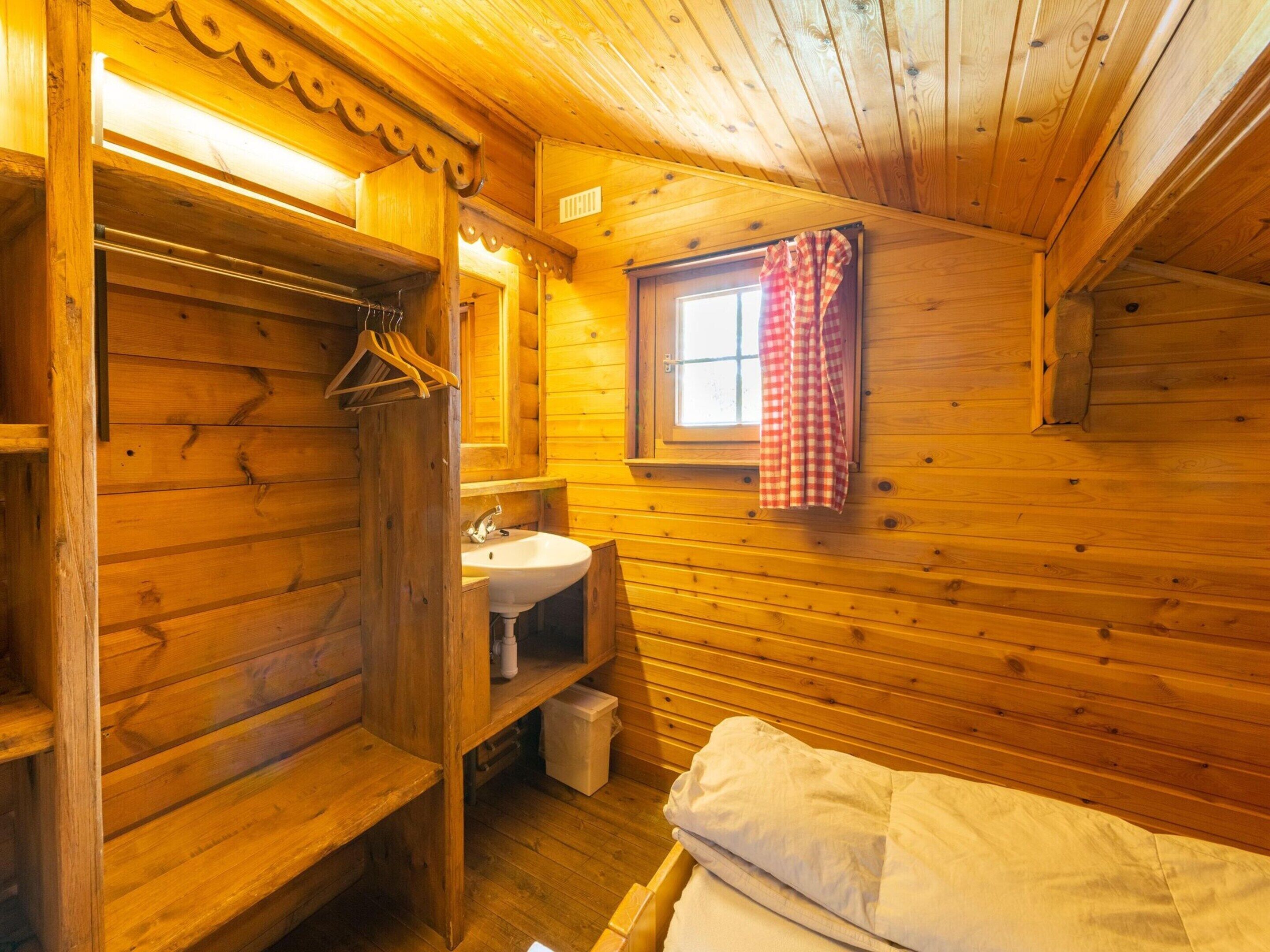L’intérieur d’une cabane en bois, avec un banc en bois, un évier, une fenêtre ornée d’un rideau à carreaux et un porte-vêtements suspendu.