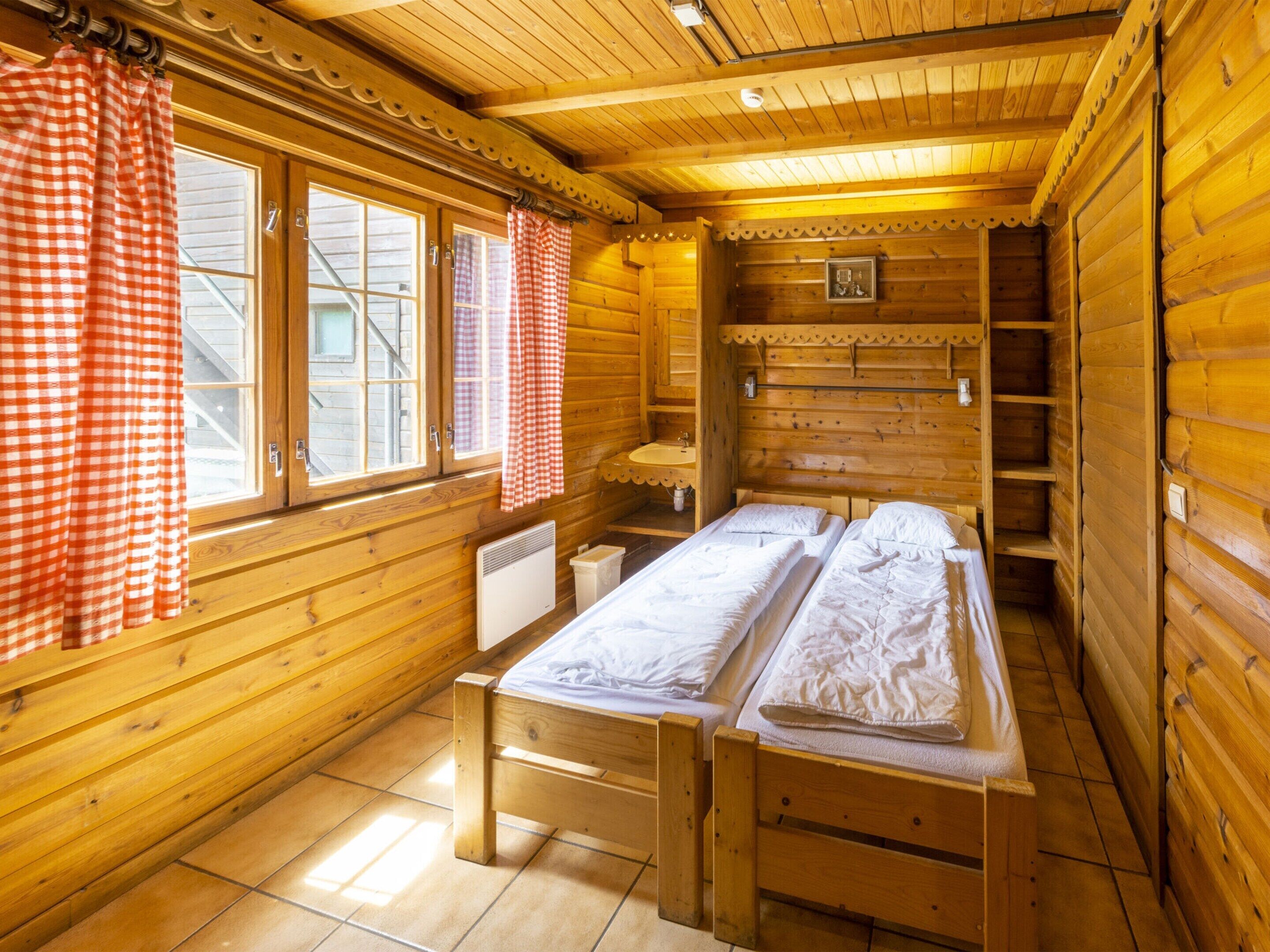 Ein Holzhütten-Zimmer mit zwei Betten, einem Fenster mit karierten Vorhängen, einem kleinen Waschbecken und einem Regal mit einem Bild.