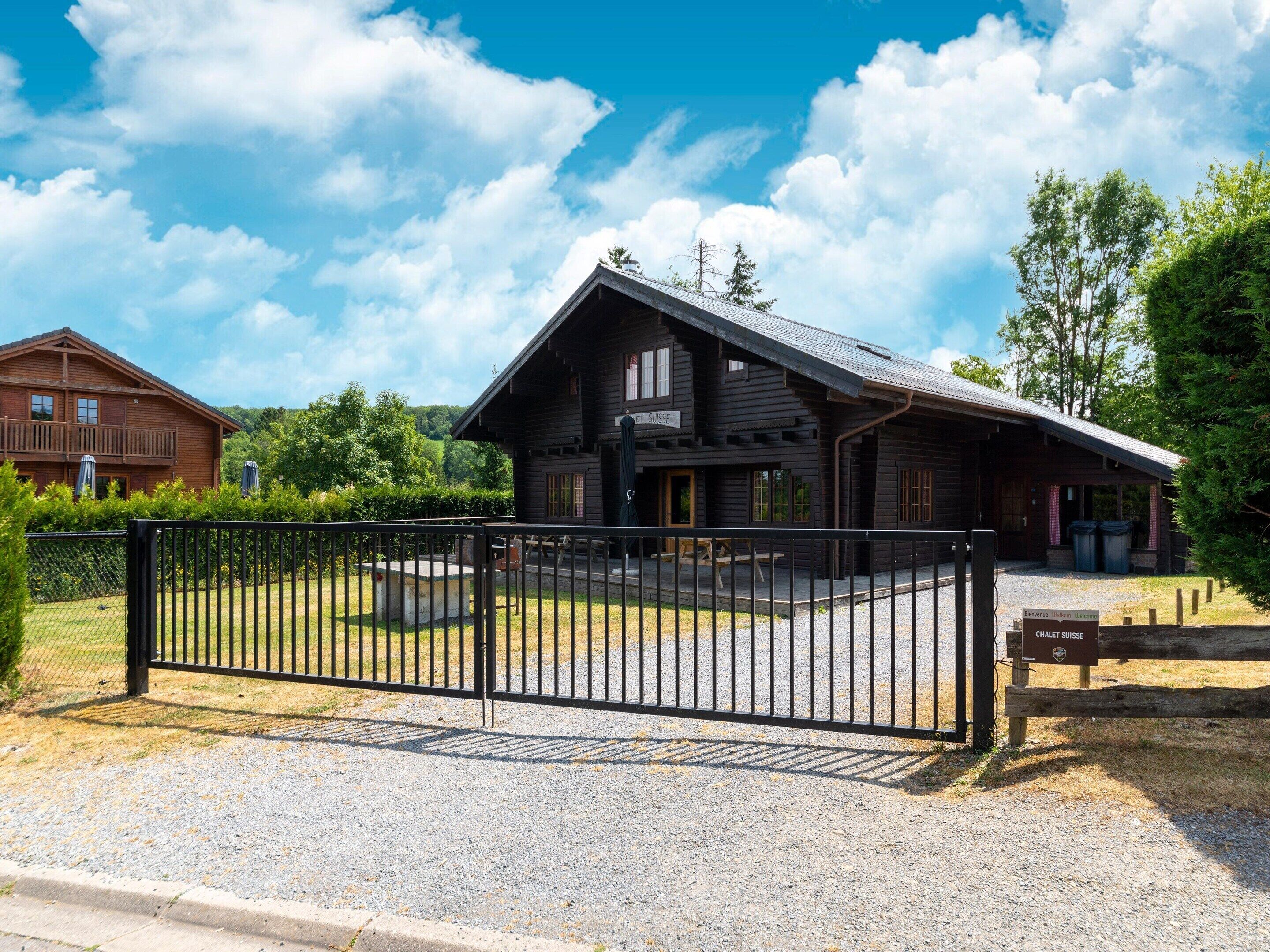 A rustic wooden chalet with a balcony, a gravel driveway, and a black metal gate.