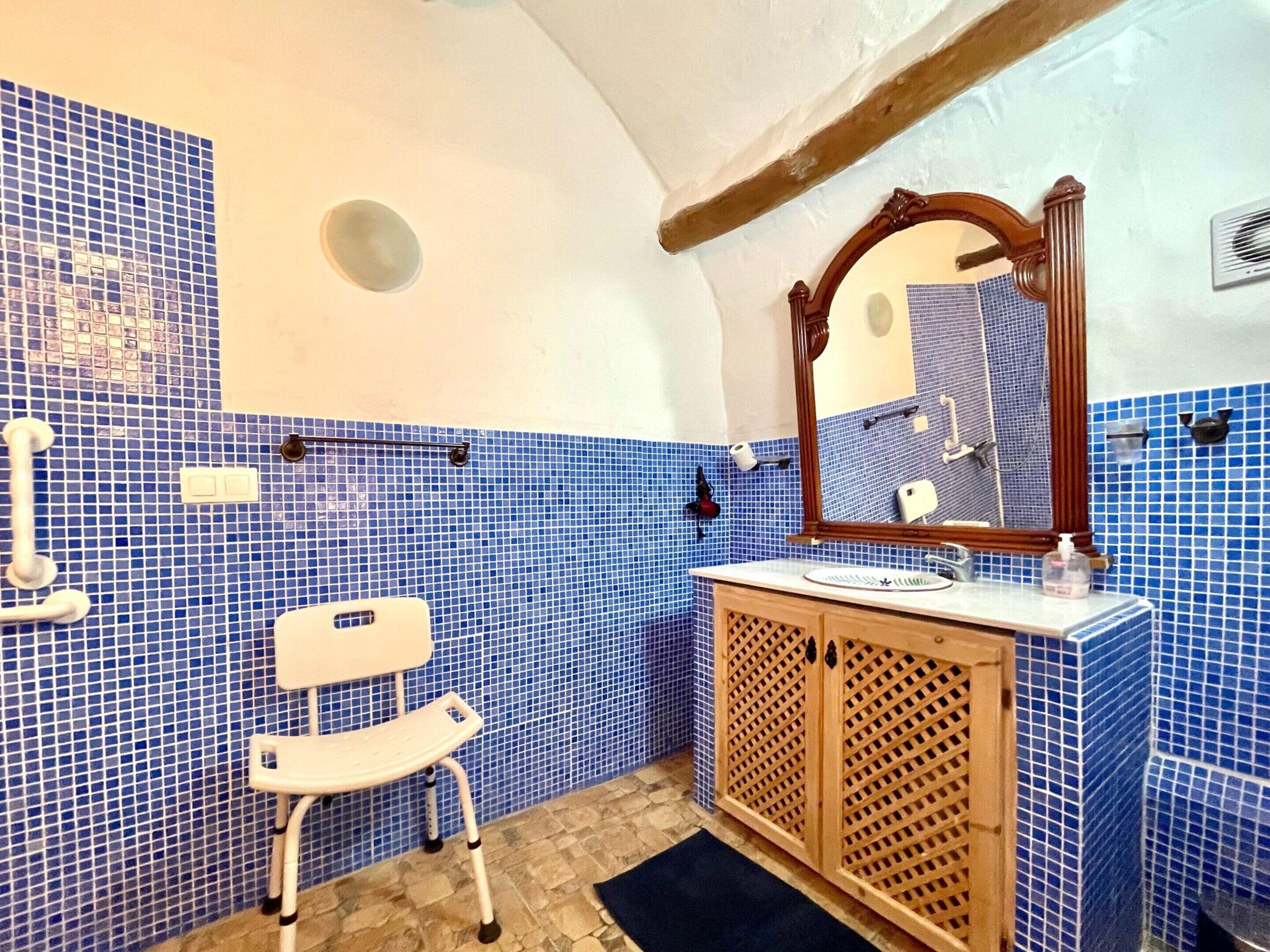 Un baño con mosaicos azules, un armario de madera, una silla blanca y un espejo.