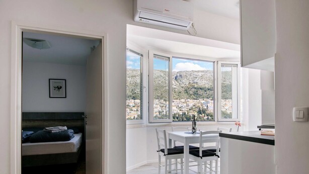 Una habitación con una cama, una zona de comedor con sillas, una mesa y vistas a un pueblo en la ladera a través de amplias ventanas.