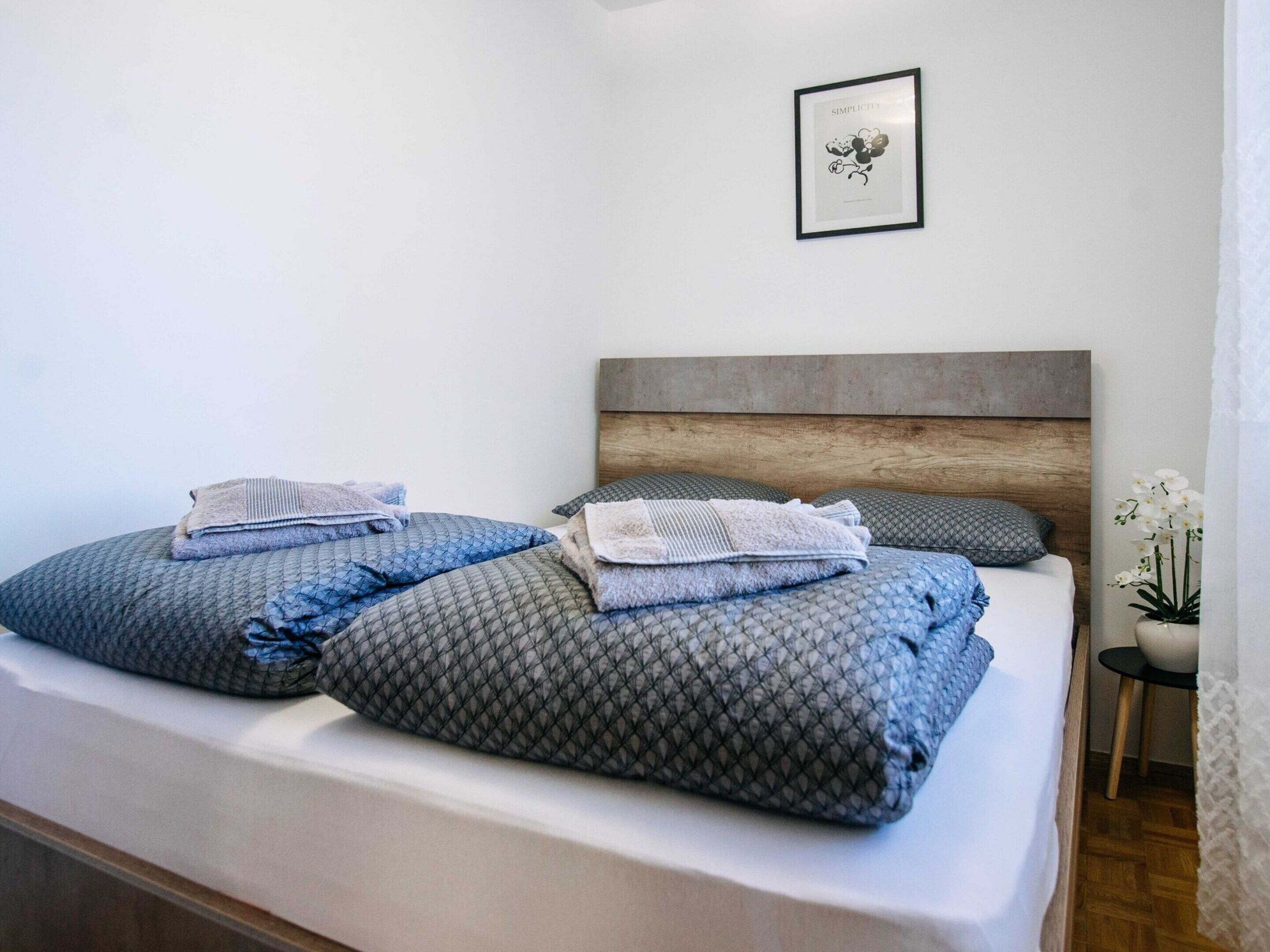 Una cama bien hecha con edredón y almohadas grises de estampado, un cabecero de madera y un cuadro en la pared.