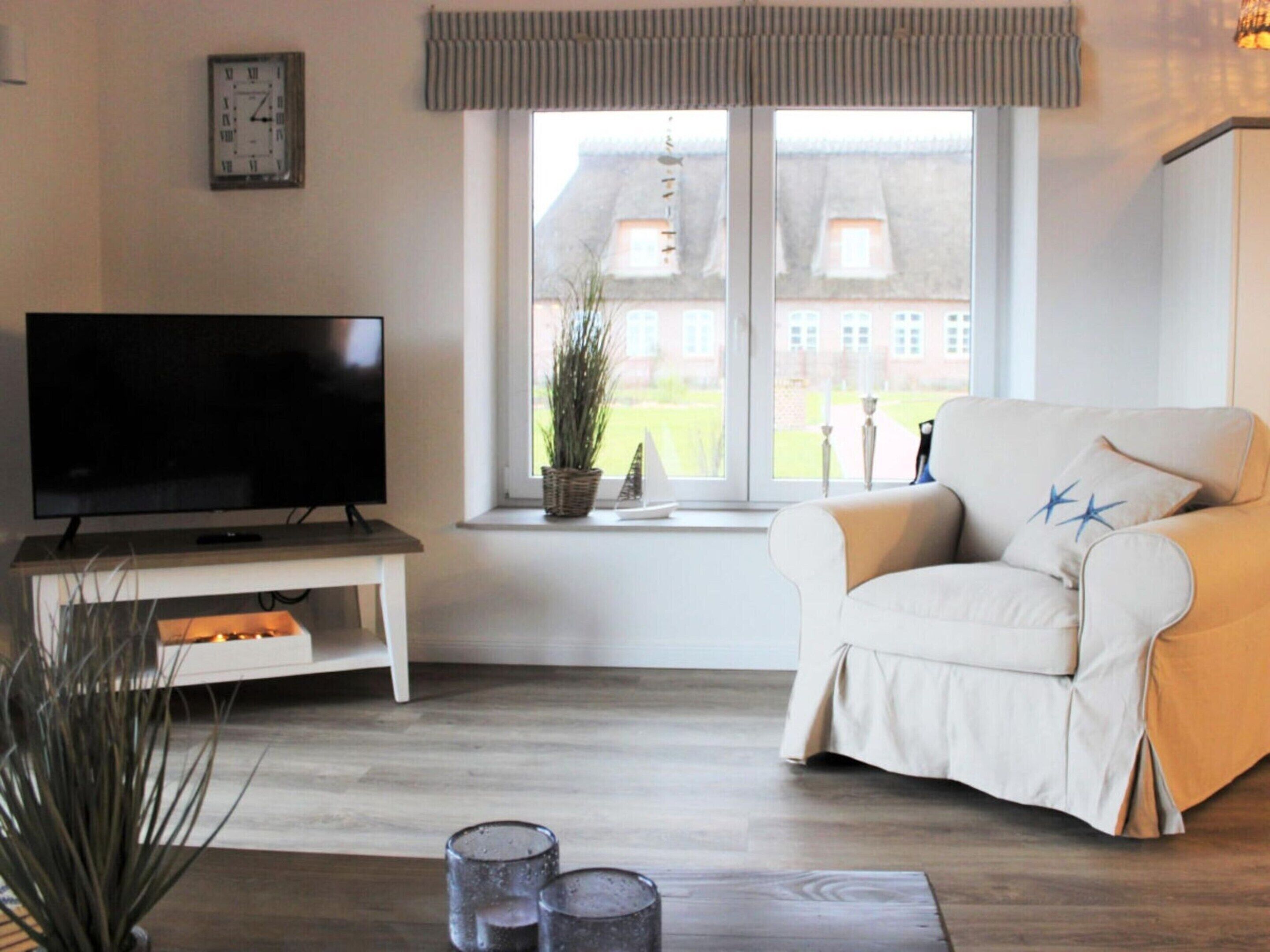 A living room with a flat-screen TV on a stand, a beige sofa with star-patterned cushions, a window with a view of buildings, and a potted plant on the windowsill.