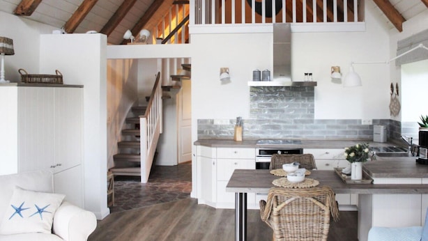Una cocina moderna con protector mural de piedra, armarios blancos y una zona de comedor con una silla de mimbre.