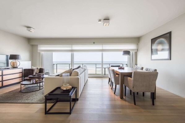 A modern living room with a sofa, coffee table, and dining area. There is a large window with a view of the beach. - Beautiful Apartment by the sea (Blankenberge)