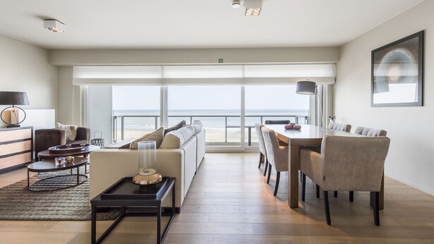 A spacious living area with a large window overlooking a beach, a dining area with a table and chairs, and a sofa with a view.