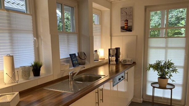 A modern kitchen with a wooden countertop, stainless steel sink, and a coffee maker. There are windows with blinds, a potted plant on a small table, and a clock on the wall. - Heimathafen Greetsiel (Krummhoern)