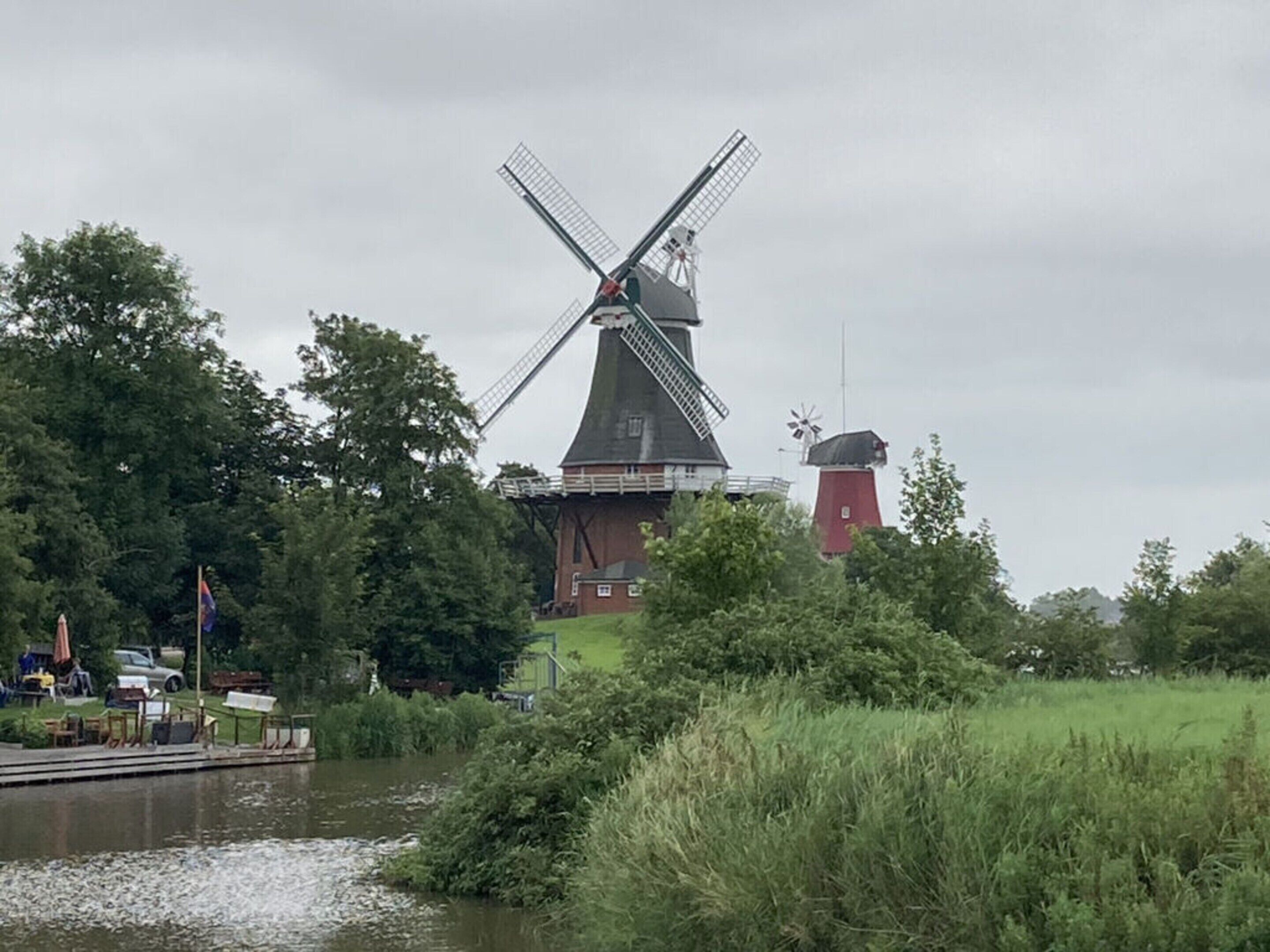 A traditional windmill with a red base and a white top, surrounded by greenery and a body of water.