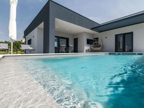 Un área de piscina moderna con agua cristalina, una zona de estar cubierta y un edificio con grandes ventanales.