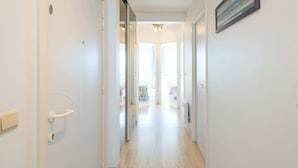 Een hal met een licht houten vloer, witte wanden en een deur met een handgreep en slot. Er hangt een ingelijst kunstwerk aan de muur en een lamp aan het plafond.