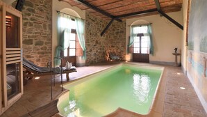 A spacious indoor swimming pool with stone walls and wooden beams, a lounge chair, and a small statue. - Belvilla by OYO Michelangelo (Castiglion Fiorentino)