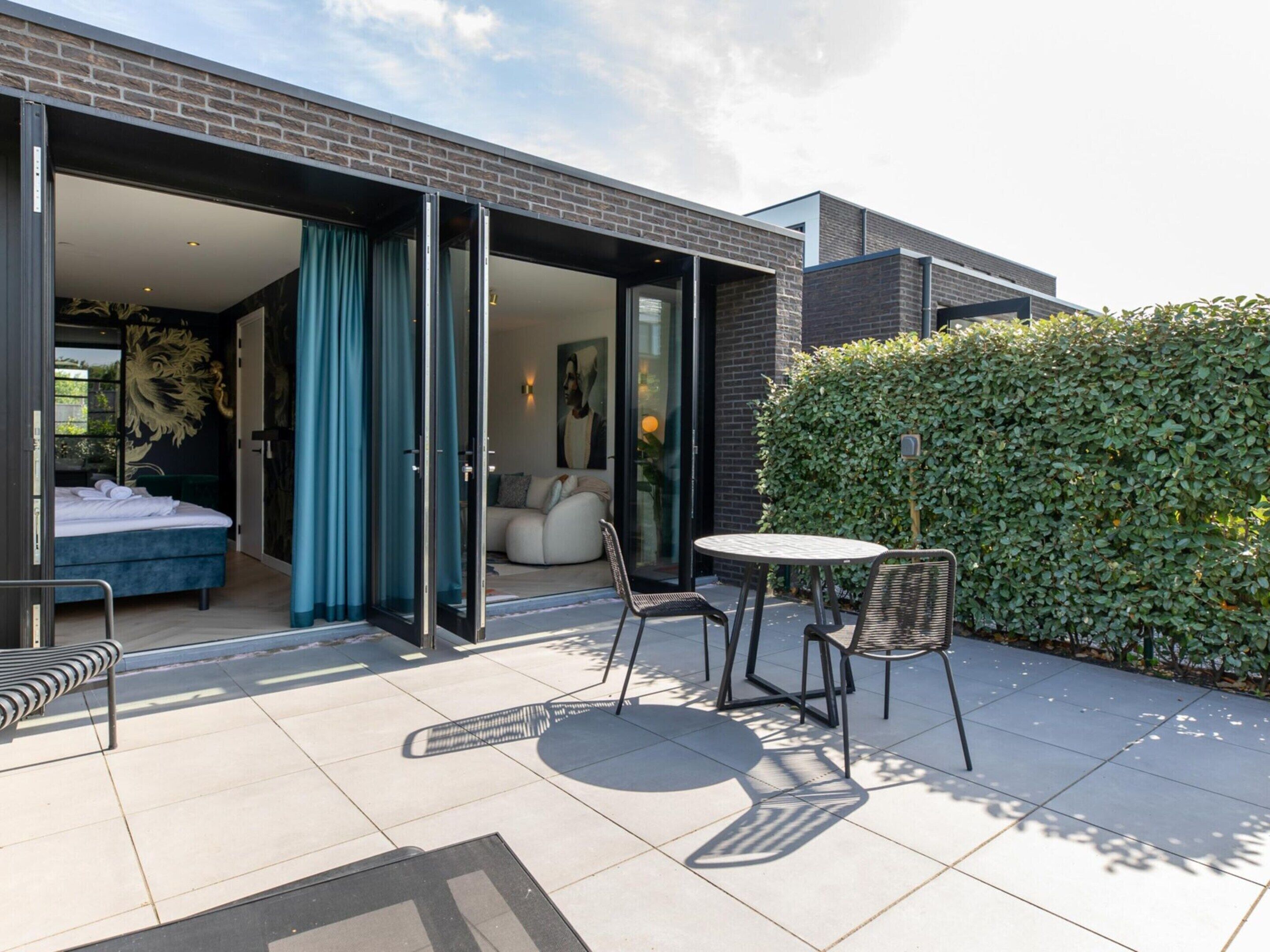 En moderne udendørs terrasse med spiseplads, en sofa og en seng, der ses gennem et åbent skydedør.