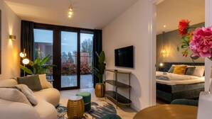 A modern hotel room with a sofa, a TV, a bed, and a balcony with a view of trees and a building. - Cozy House in Koudekerke Near the Beach (Koudekerke)