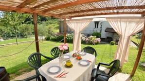 Ein Essbereich im Freien mit einem Tisch für vier Personen, umgeben von Stühlen, unter einer Holzw pergola mit weißem Tischleinentuch.