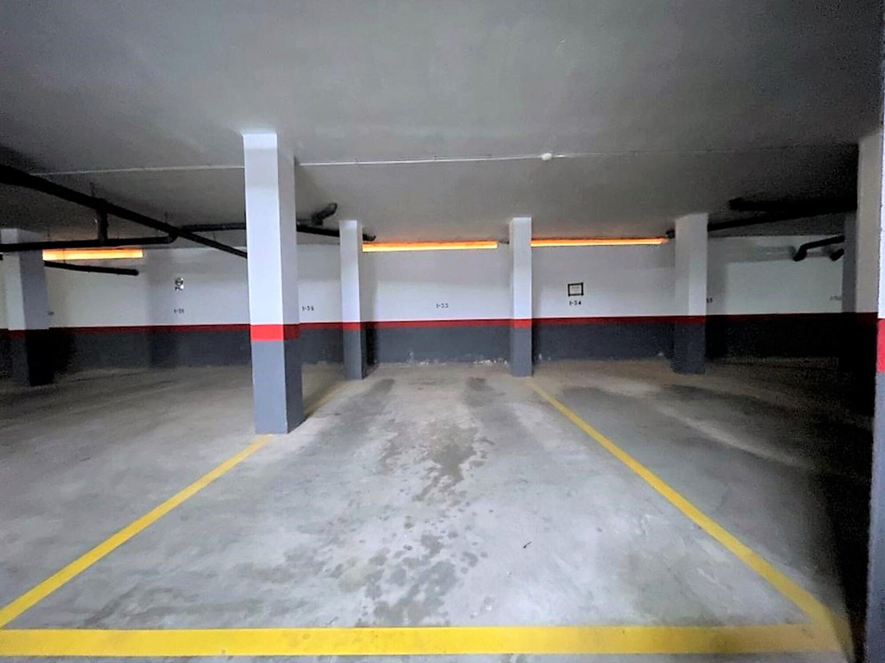 Un estacionamiento interior con suelo y columnas de hormigón, plazas delimitadas y iluminación en el techo.