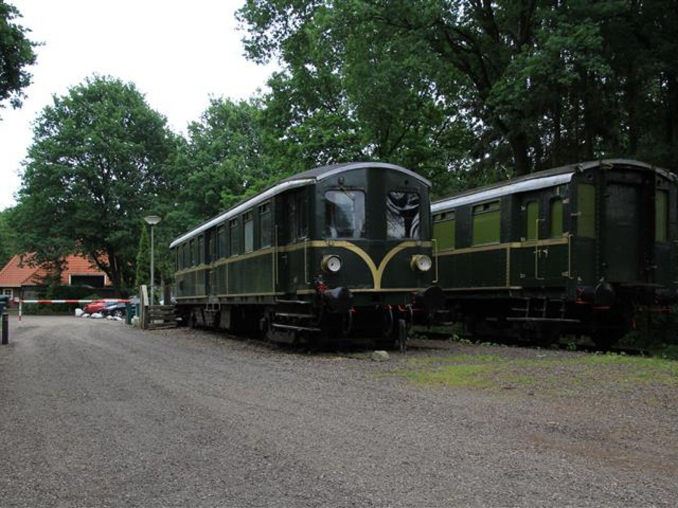 A vintage train parked on a gravel track with trees and a building in the background.