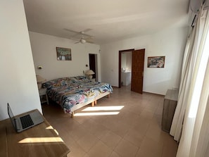 Une chambre à coucher comprenant un lit, un bureau avec un ordinateur portable, un ventilateur de plafond et deux portes donnant sur d’autres pièces.