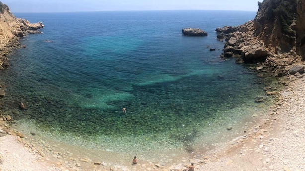 Une plage côtière à l’eau claire, avec un terrain rocheux et une vue lointaine sur la mer.