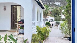 Un bâtiment blanc avec une entrée carrelée et un balcon. Un sentier mène à une colline, avec des maisons visibles au loin.