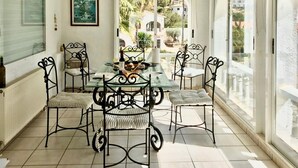 Une terrasse ensoleillée avec une table en verre entourée de chaises en métal, un tableau encadré au mur et une vue sur la verdure à l’extérieur.