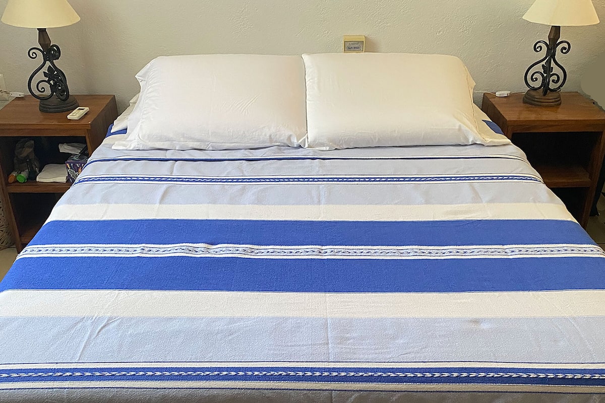Un lit avec une couvre-lit à rayures, deux tables de chevet avec des lampes et une petite étagère avec quelques objets posés sur l’une des tables.