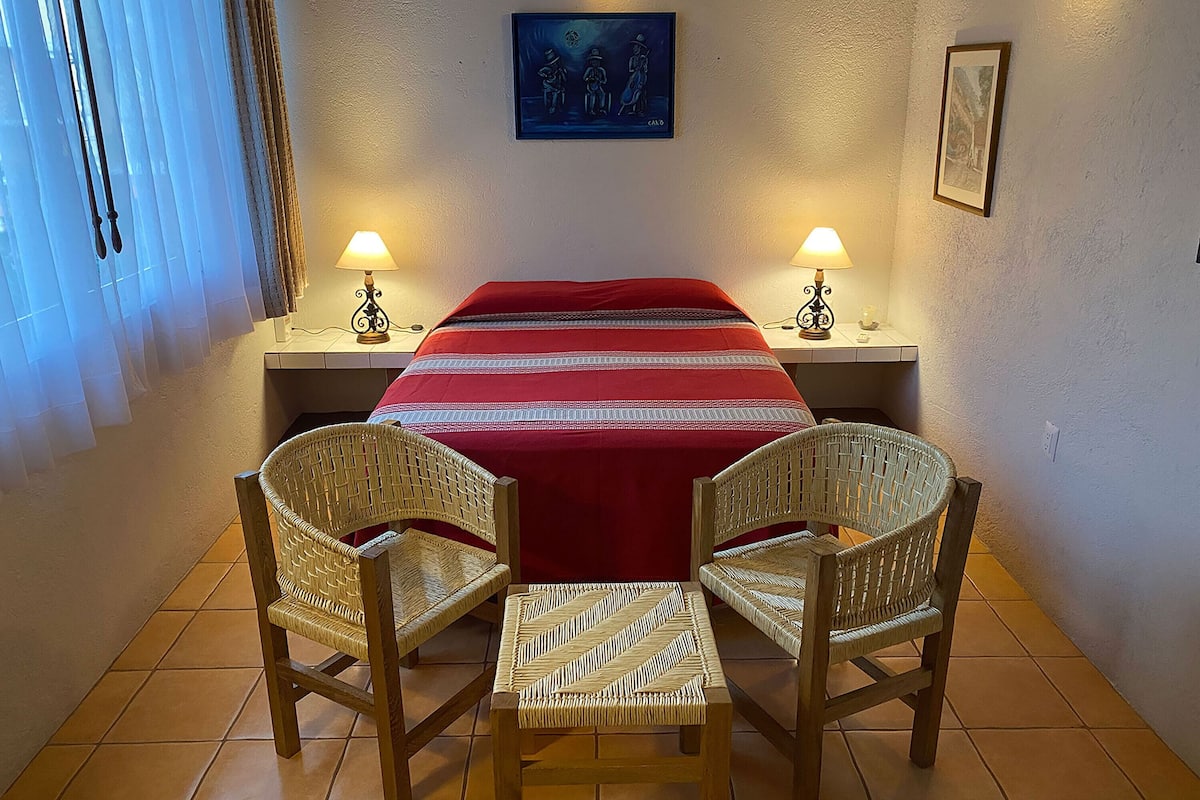 Une chambre d’hôtel avec un lit, deux fauteuils en osier, une couvre-lit rouge, deux lampes fixées au mur et un tableau encadré accroché au mur.