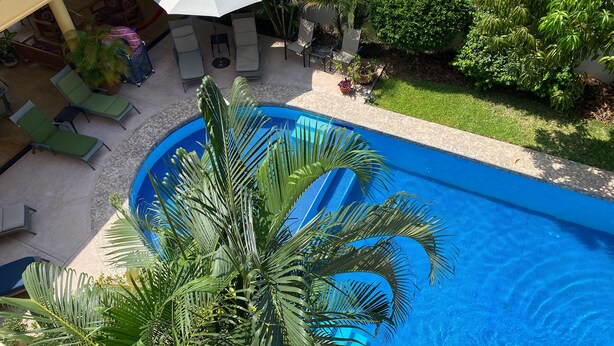 Una piscina con sillas de descanso y sombrillas, rodeada de vegetación y una zona de estar cubierta.