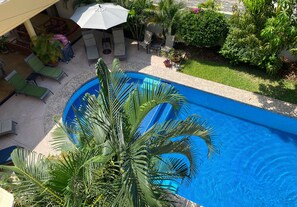 Una piscina con sillas de descanso y sombrillas, rodeada de vegetación y una zona de estar cubierta.